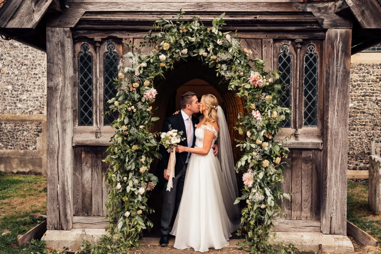 newlywed couple standing in a church doorway under flowery arch kissing whikst being photographed by Wedding Photographer based in Northamptonshire.