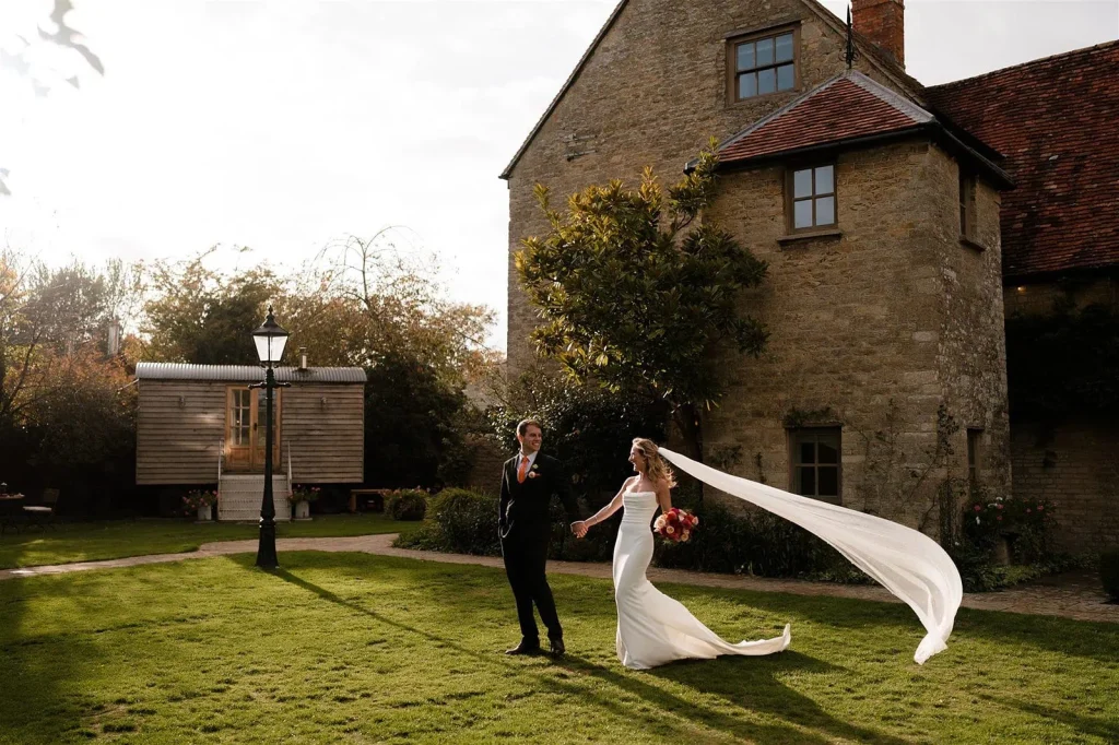 couple walking on the grounds of Tythe barn, brides vail is being wind swept whilst being photographed by Oxfordshire documentary wedding photographer